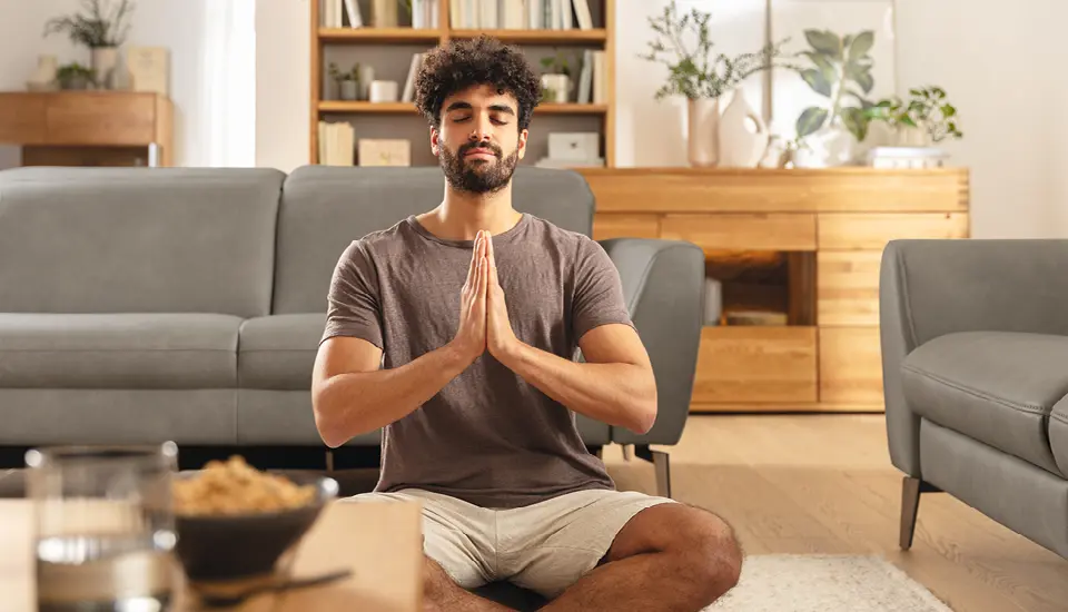 Mann sitzt im Schneidersitz auf einem hellen Teppich vor einem grauen Sofa und meditiert, im Hintergrund Sideboard und Bücherregal aus hellem Holz, üppige Pflanzen und warmes Tageslicht  